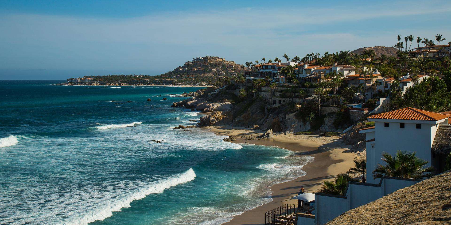 cabo-san-lucas-pacific-coast A stunning coastal scene of Cabo San Lucas featuring turquoise Pacific waters with white-capped waves rolling onto a sandy beach, framed by luxurious white and terracotta-roofed villas perched on rocky cliffs. Palm trees dot the hillside where additional residential properties cascade down to meet the pristine shoreline.