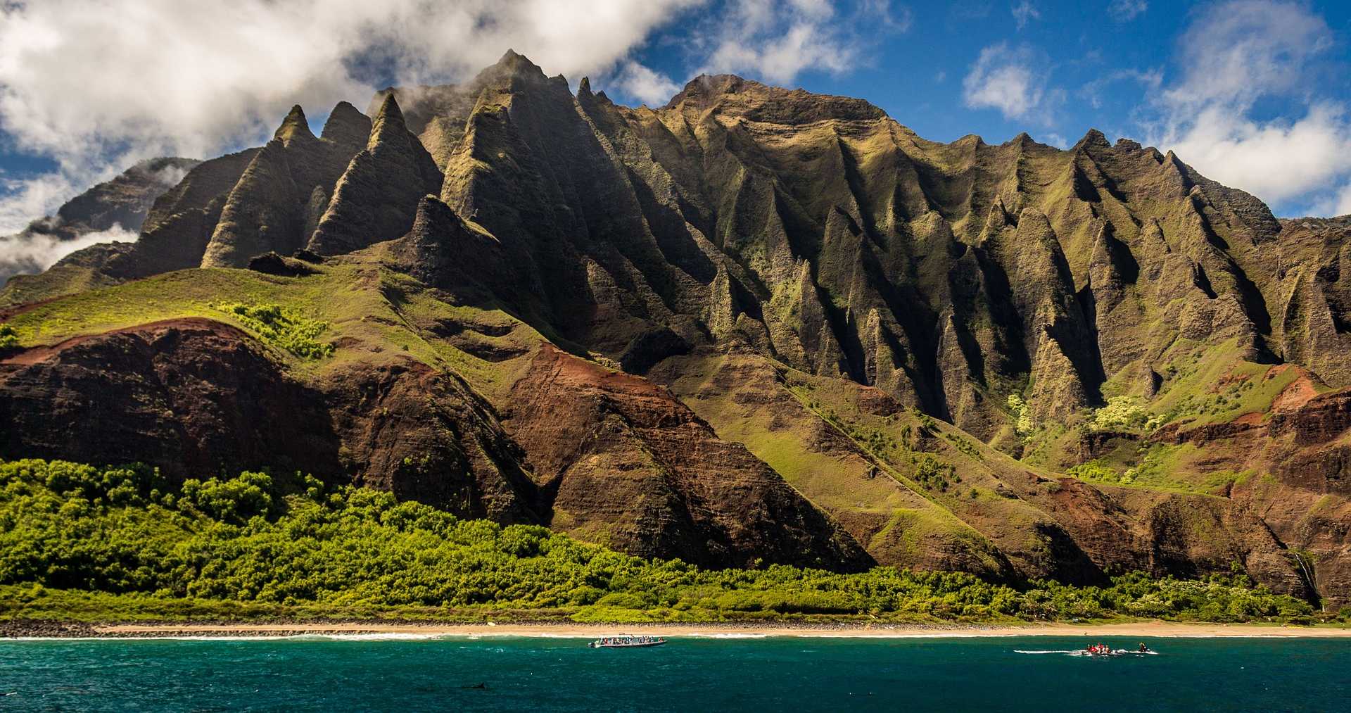 hawaii-pacific Towering verdant sea cliffs rise dramatically from a turquoise ocean in this breathtaking view of Kalalau Valley on Kauai's remote Na Pali Coast. Lush green vegetation blankets the valley floor and lower slopes, contrasting sharply with the jagged, erosion-carved ridges of the cliff face and anchored boats offshore.