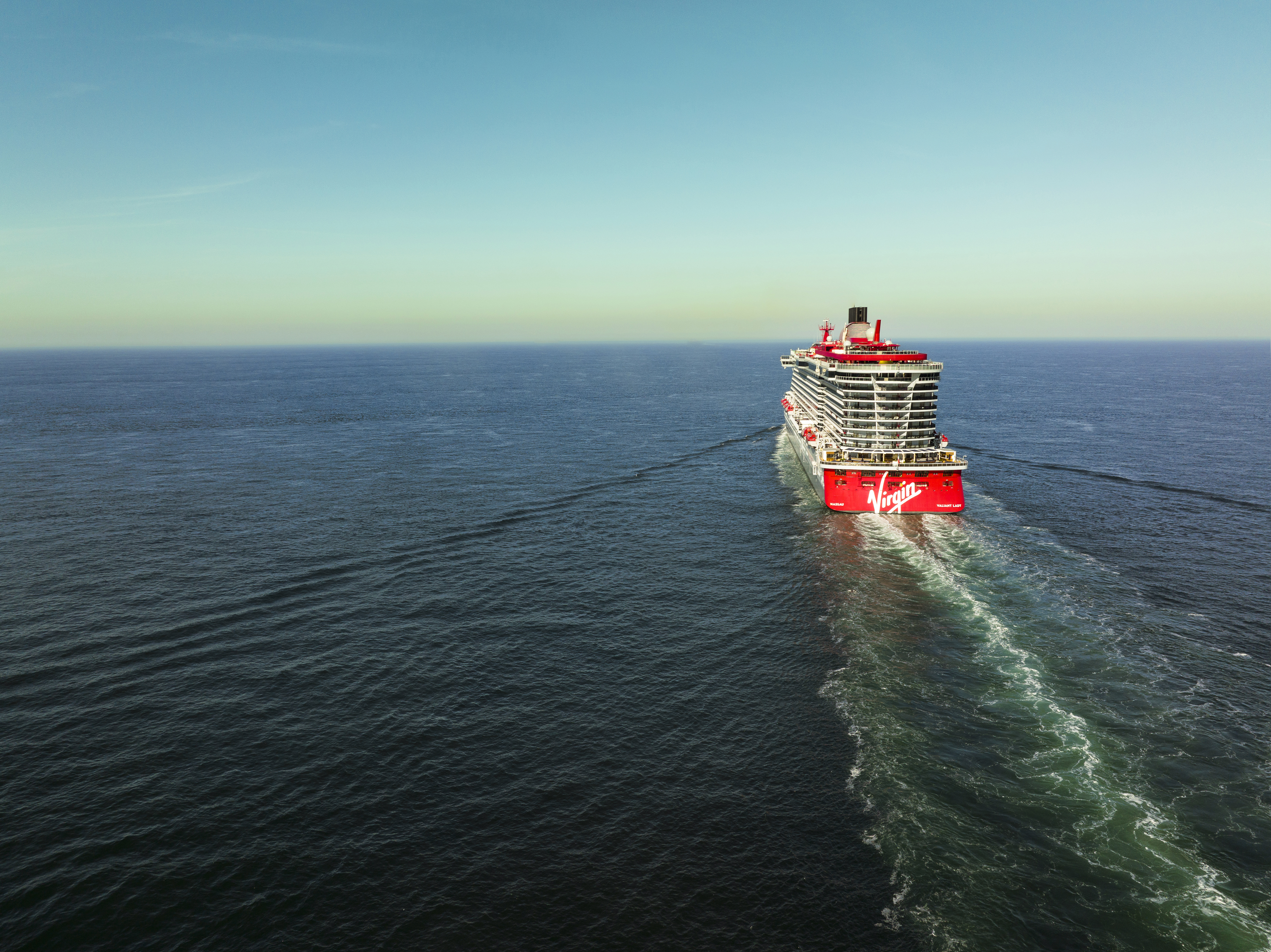 footer-image-Valiant-Lady-Exterior-Sailing-Far-Away A striking aerial view of the Virgin Voyages' Valiant Lady cruise ship sailing through calm ocean waters, with its distinctive red hull and modern multi-deck design clearly visible against a serene blue sea and clear sky.