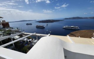 Scenic view of Santorini, Greece, overlooking the deep blue Aegean Sea with several cruise ships anchored near volcanic islands, whitewashed terrace restaurants in the foreground, and rugged cliffs in the distance under a bright blue sky with scattered clouds.