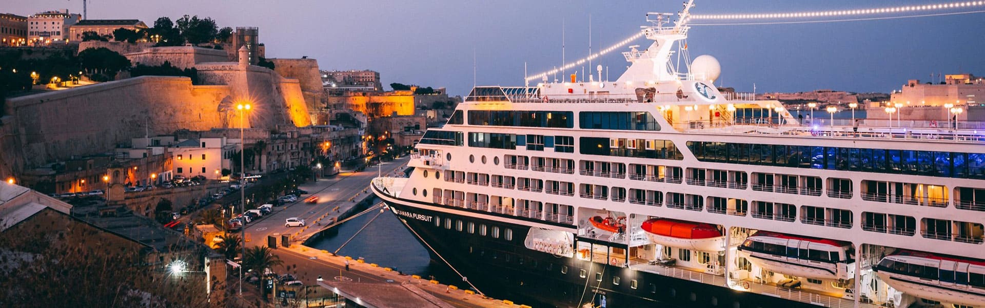 A modern luxury cruise ship is moored in the historic Grand Harbour of Valletta, Malta, with its illuminated white hull contrasting against the warmly-lit fortified walls and buildings of the UNESCO World Heritage city at twilight. The ship's multiple decks and contemporary design showcase the scale of modern cruise vessels alongside centuries-old Mediterranean architecture.
