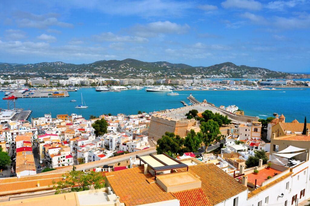 A panoramic view of Ibiza Town in the Balearic Islands, Spain, with whitewashed houses and terracotta rooftops overlooking a bright blue harbor filled with yachts and ferries, backed by green hills under a sunny sky.