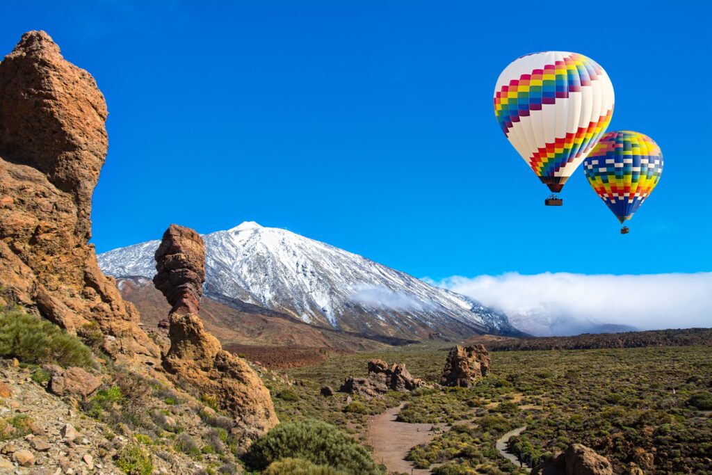 Colorful hot air balloons floating over the volcanic landscape of Mount Teide in Tenerife, Canary Islands, with snow-capped peaks, rugged rock formations, and clear blue skies.