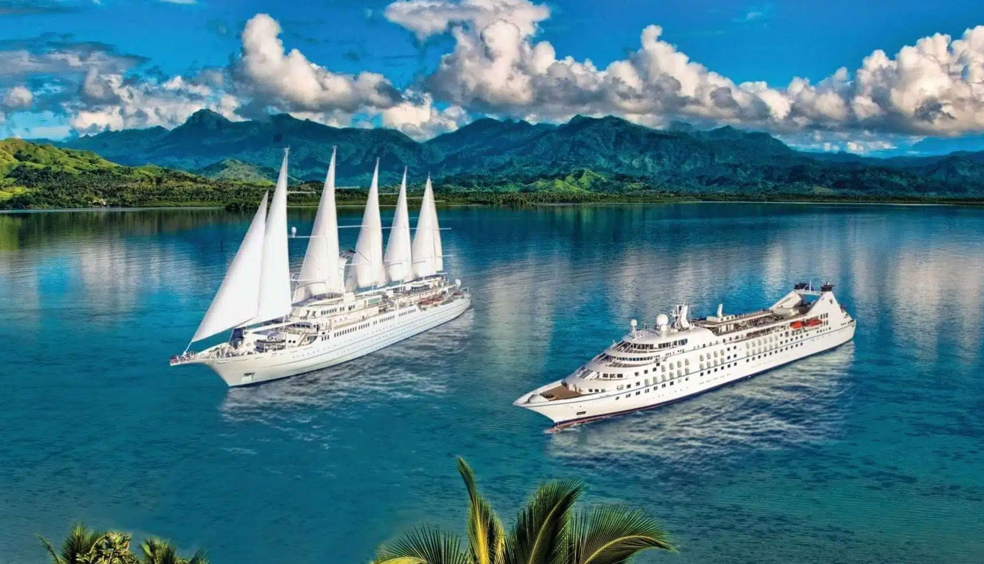 Two elegant cruise vessels anchored in crystal-clear turquoise waters with lush, forested mountains and dramatic clouds in the background. The left ship features distinctive white sails, while the right vessel is a modern motor cruise ship. Palm fronds are visible in the foreground, suggesting a tropical Pacific destination.