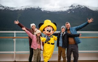 A smiling family poses on deck with a costumed Disney character in bright yellow rain gear during an Alaskan cruise, snow-capped mountains rising in the background.