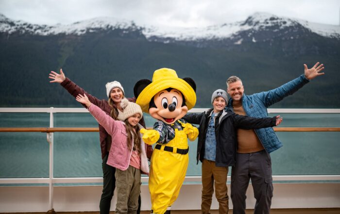 A smiling family poses on deck with a costumed Disney character in bright yellow rain gear during an Alaskan cruise, snow-capped mountains rising in the background.