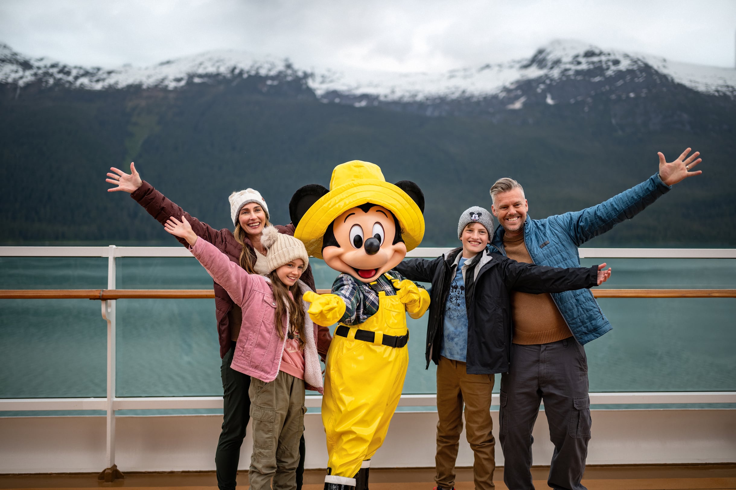 A smiling family poses on deck with a costumed Disney character in bright yellow rain gear during an Alaskan cruise, snow-capped mountains rising in the background.
