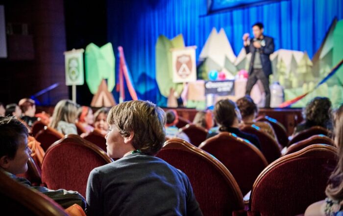 Children sit in a cruise ship theater watching a lively educational presentation with colorful forest-themed props as part of the Discovery at SEA program.