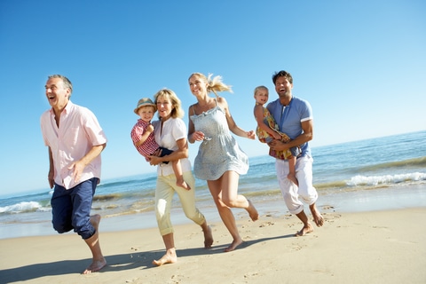 Three generations of a family run joyfully along a sunny beach, laughing and holding hands by the shoreline.