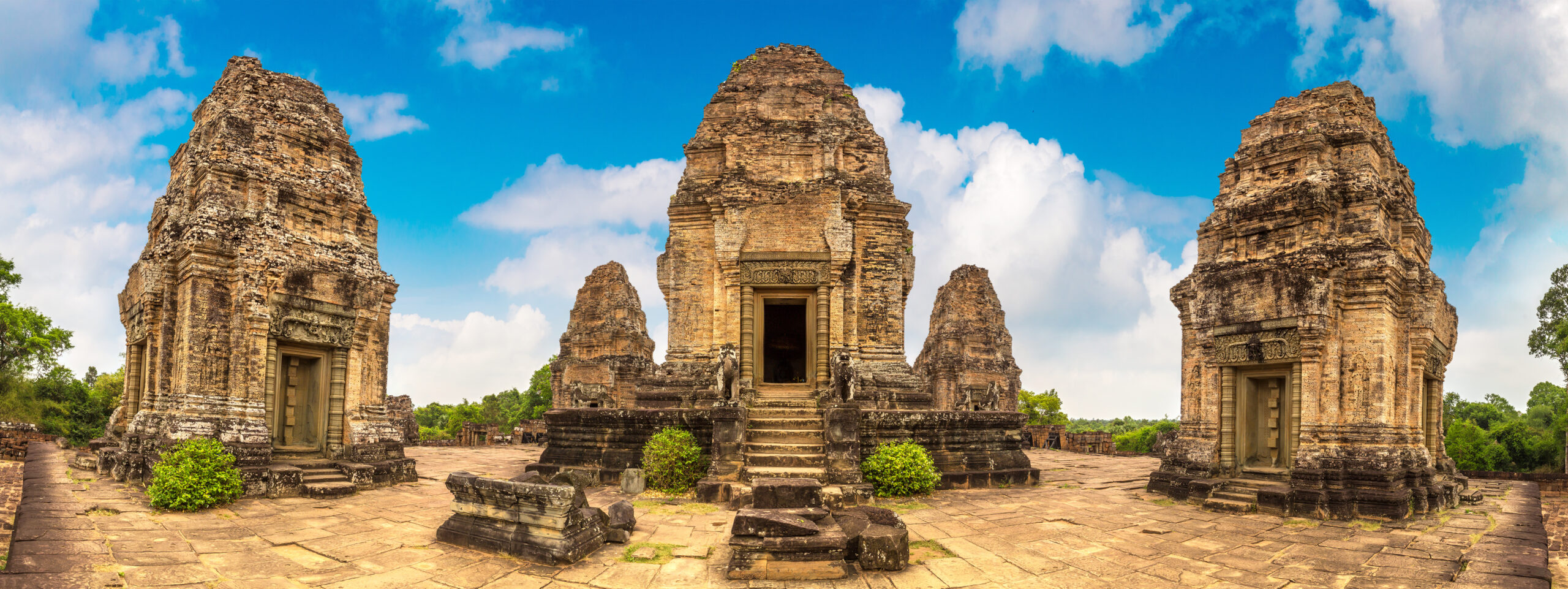 Ancient Khmer Temples of Angkor Archaeological Site A panoramic view of three large stone temple towers with intricate architectural details, located in a historic Khmer temple complex with a bright blue sky and scattered clouds.