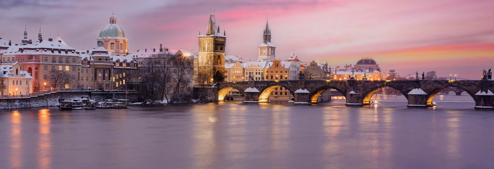 A stunning winter view of the historic Charles Bridge spanning the Vltava River in Prague, Czech Republic, with illuminated baroque buildings and a pastel pink and purple sunset sky.