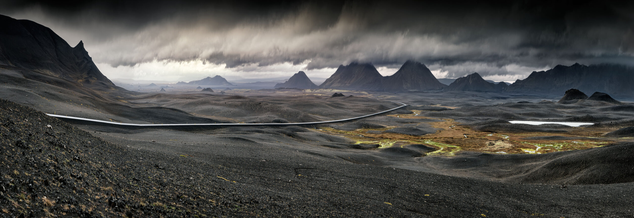 A stark, moody landscape featuring dark volcanic terrain with a winding road cutting through barren plains, surrounded by rugged mountain peaks shrouded in dramatic storm clouds.