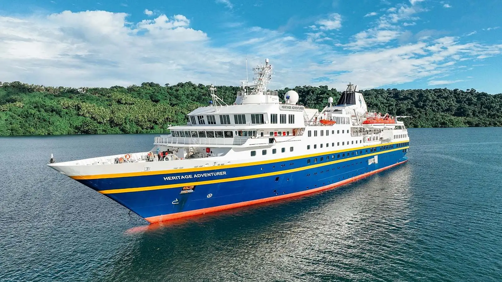 Heritage Adventurer Expedition Cruise Ship The Heritage Adventurer, a modern expedition cruise ship with distinctive blue and orange hull livery, anchors in turquoise tropical waters surrounded by lush green mangrove forests. The vessel features multiple decks, advanced navigation equipment, and is designed for adventure cruising to remote destinations.