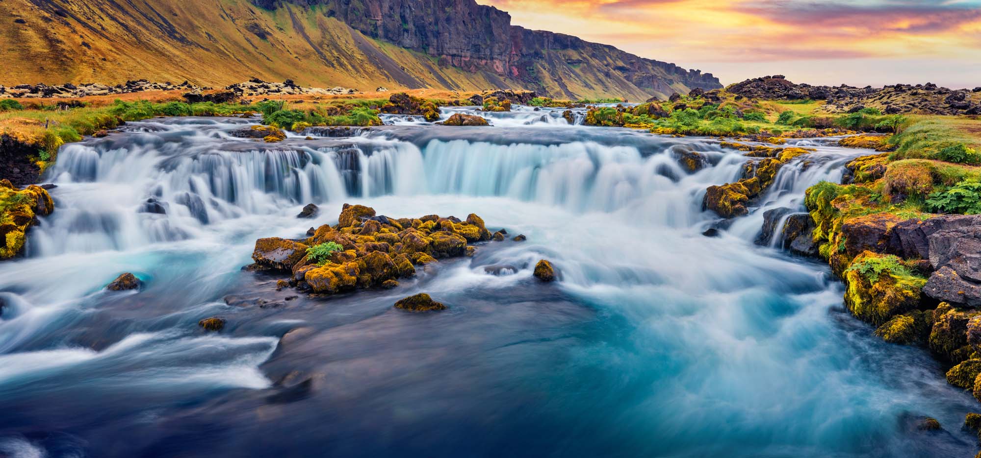 A stunning multi-tiered waterfall cascading over moss-covered rocks with towering basalt cliffs in the background. The scene features flowing turquoise water, golden-hued rocks, and a dramatic sunset sky.