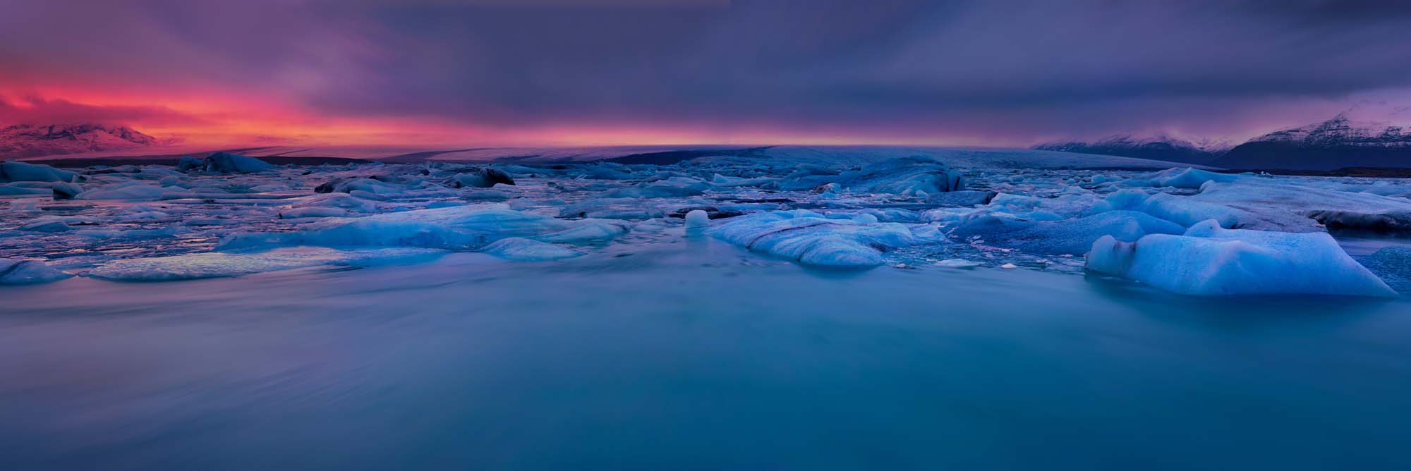 A breathtaking panorama of floating blue icebergs in a tranquil glacial lagoon, with a dramatic pink and purple sunset illuminating snow-capped mountains in the background.