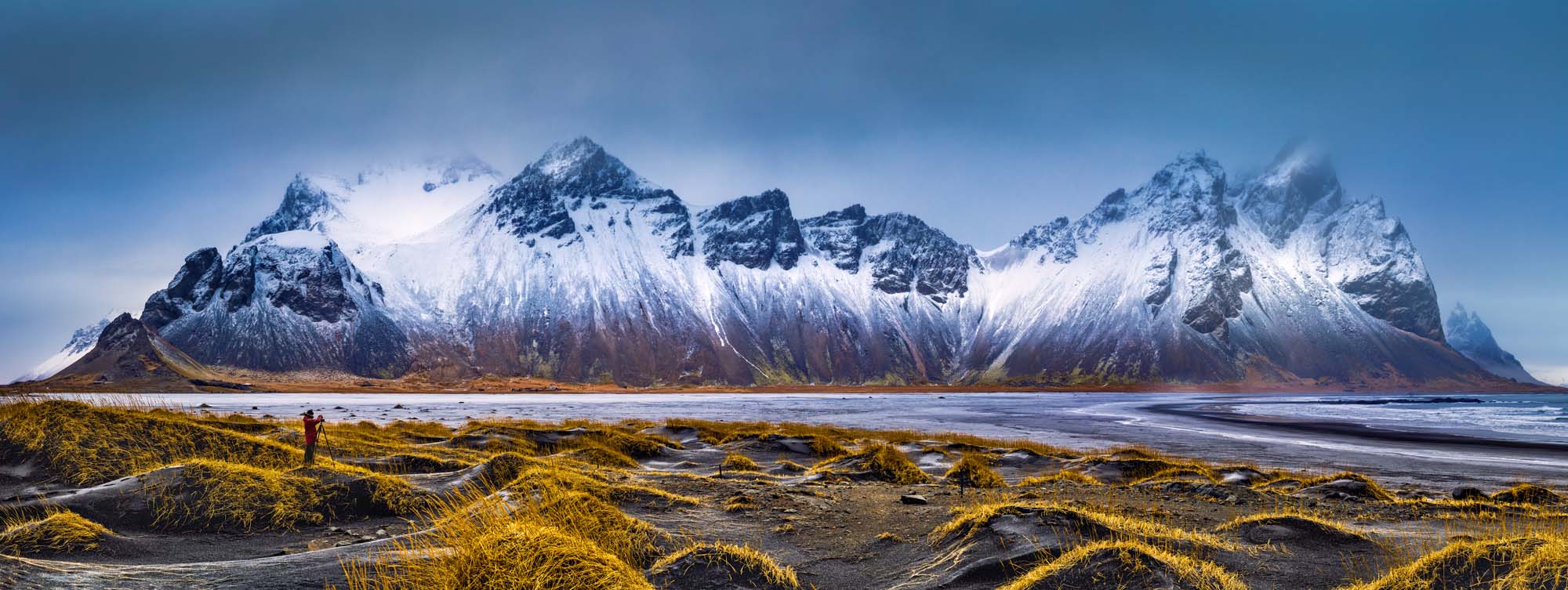 A dramatic landscape featuring rugged, snow-covered mountains with sharp peaks against a blue sky, overlooking a black sand beach with golden grass in the foreground. A solitary figure stands in the landscape, providing scale to the immense natural scenery.