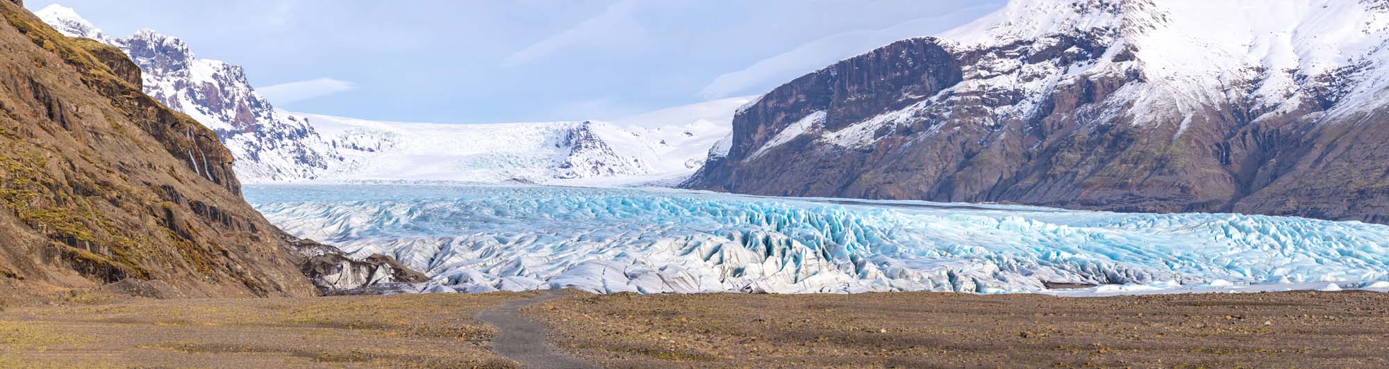 A vast blue-white glacier stretching between steep, rocky mountain slopes with snow-capped peaks. The glacier's textured surface displays intricate patterns of ice in varying shades of blue and white.