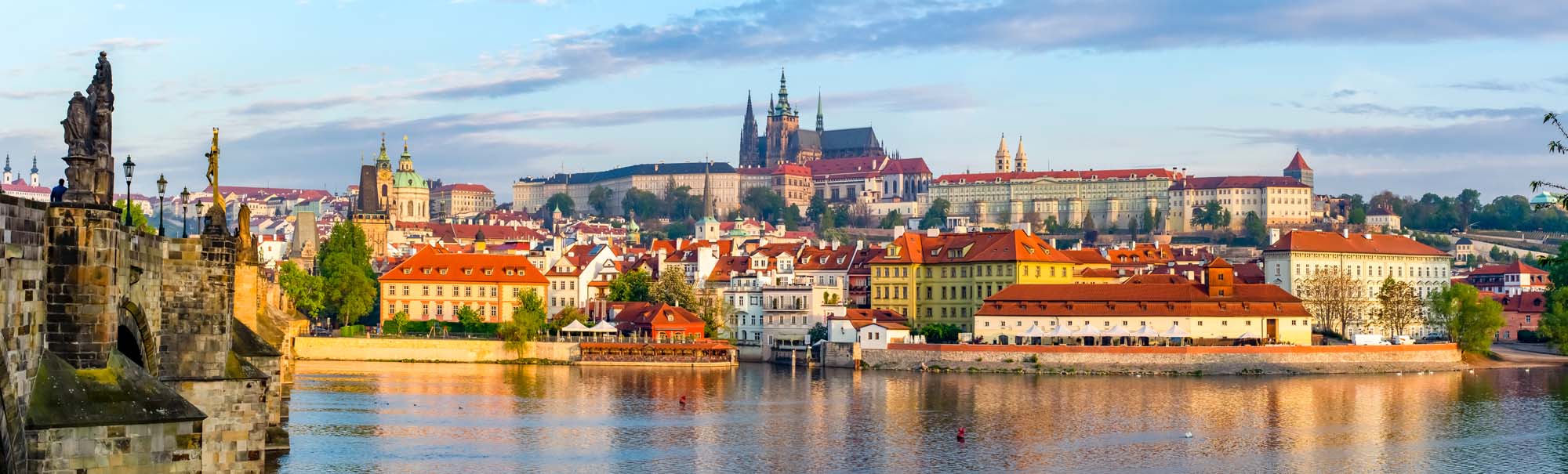 A stunning panoramic view of Prague's historic city center, featuring the Prague Castle complex, colorful baroque buildings, and the Vltava River with the iconic Charles Bridge in the foreground.