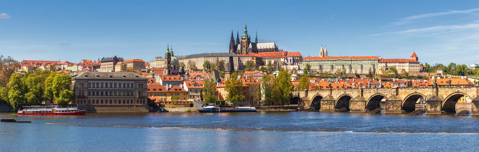 A stunning panoramic view of Prague's historic cityscape featuring Prague Castle, the Charles Bridge, and the Vltava River under a clear blue sky.