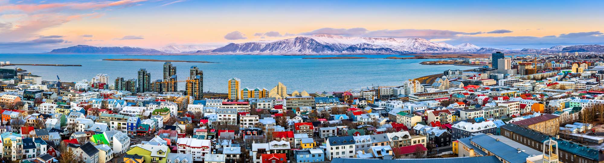 Reykjavik, Iceland – Colorful City Panorama A vibrant aerial view of Reykjavik showcasing colorful rooftops, modern high-rise buildings, and snow-capped mountains in the background with a serene ocean view.