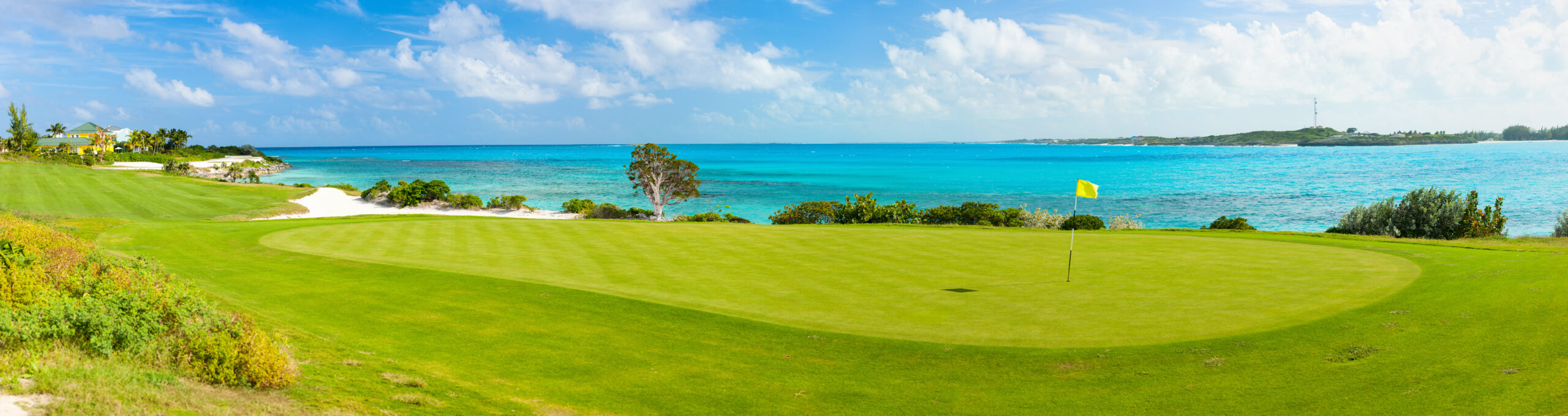 Tropical Oceanfront Golf Course A pristine golf green overlooking turquoise waters with a yellow flag and lush landscaping. The course features perfectly manicured grass with a stunning ocean and island backdrop.
