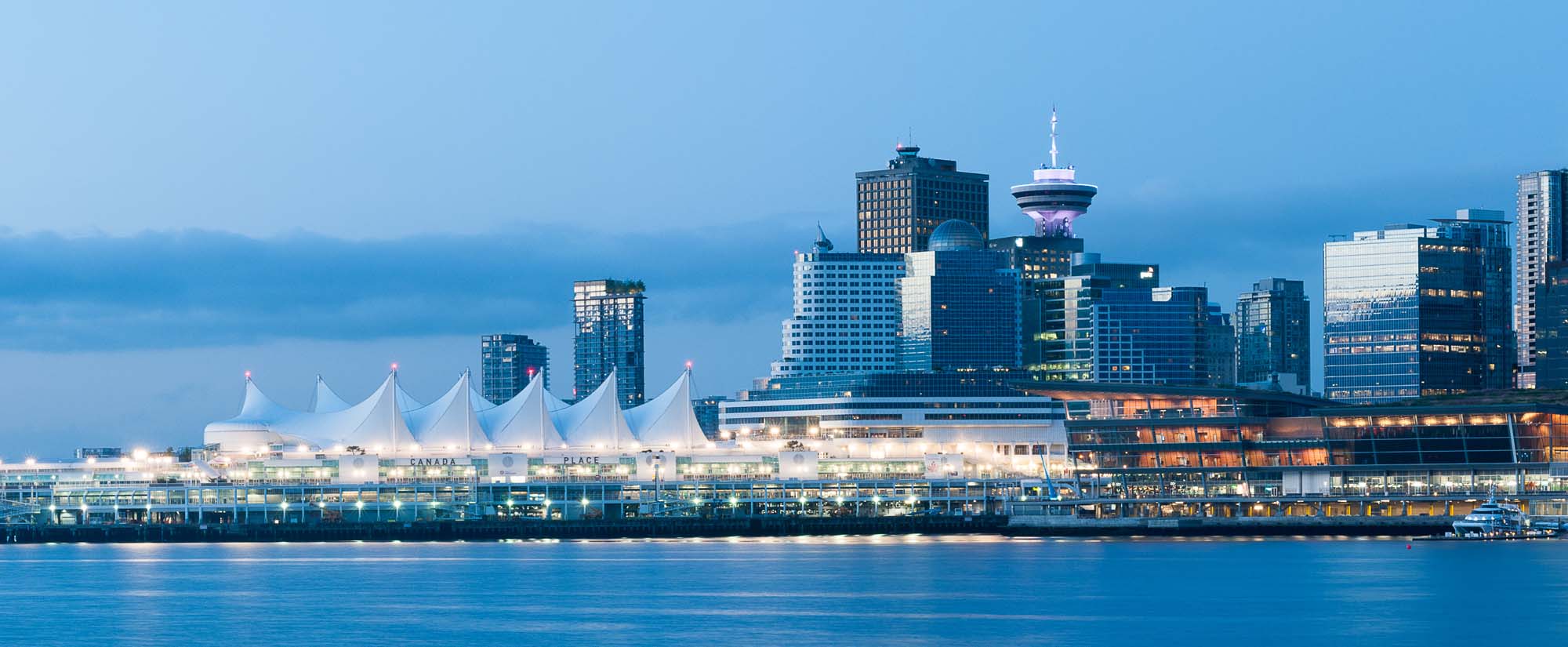 A panoramic view of Vancouver's Canada Place cruise terminal and downtown skyline at dusk, featuring modern architectural buildings and a scenic waterfront setting.