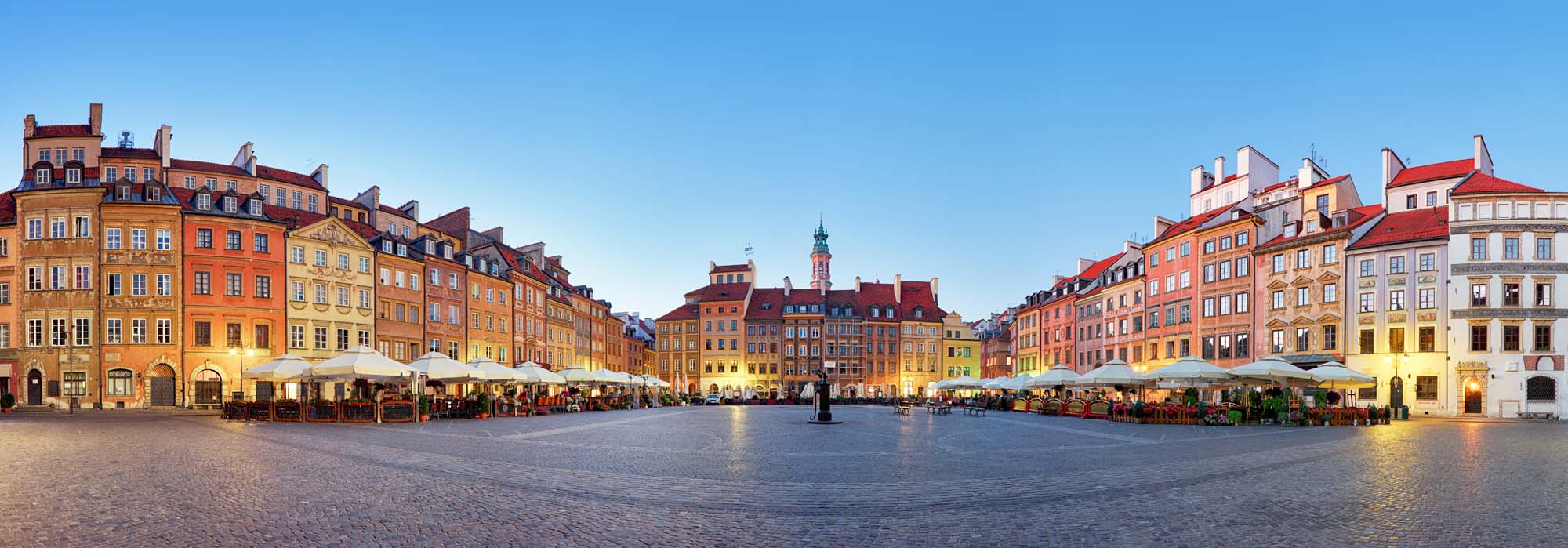 A picturesque panoramic view of the historic Old Town Market Square in Warsaw, Poland, featuring colorful baroque townhouses and outdoor cafe terraces at twilight.
