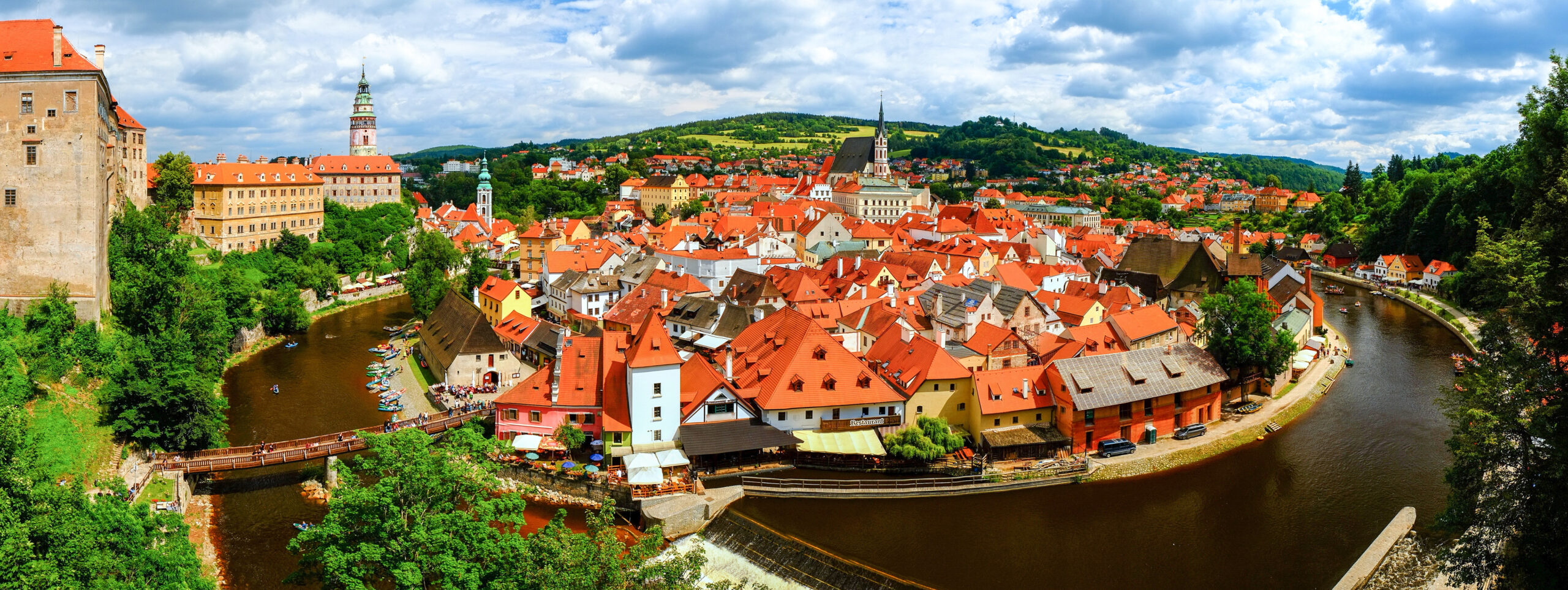 A picturesque aerial view of Český Krumlov, a UNESCO World Heritage Site in the Czech Republic, featuring tightly clustered red-roofed buildings nestled along a meandering river with a castle and church towers rising above the landscape.