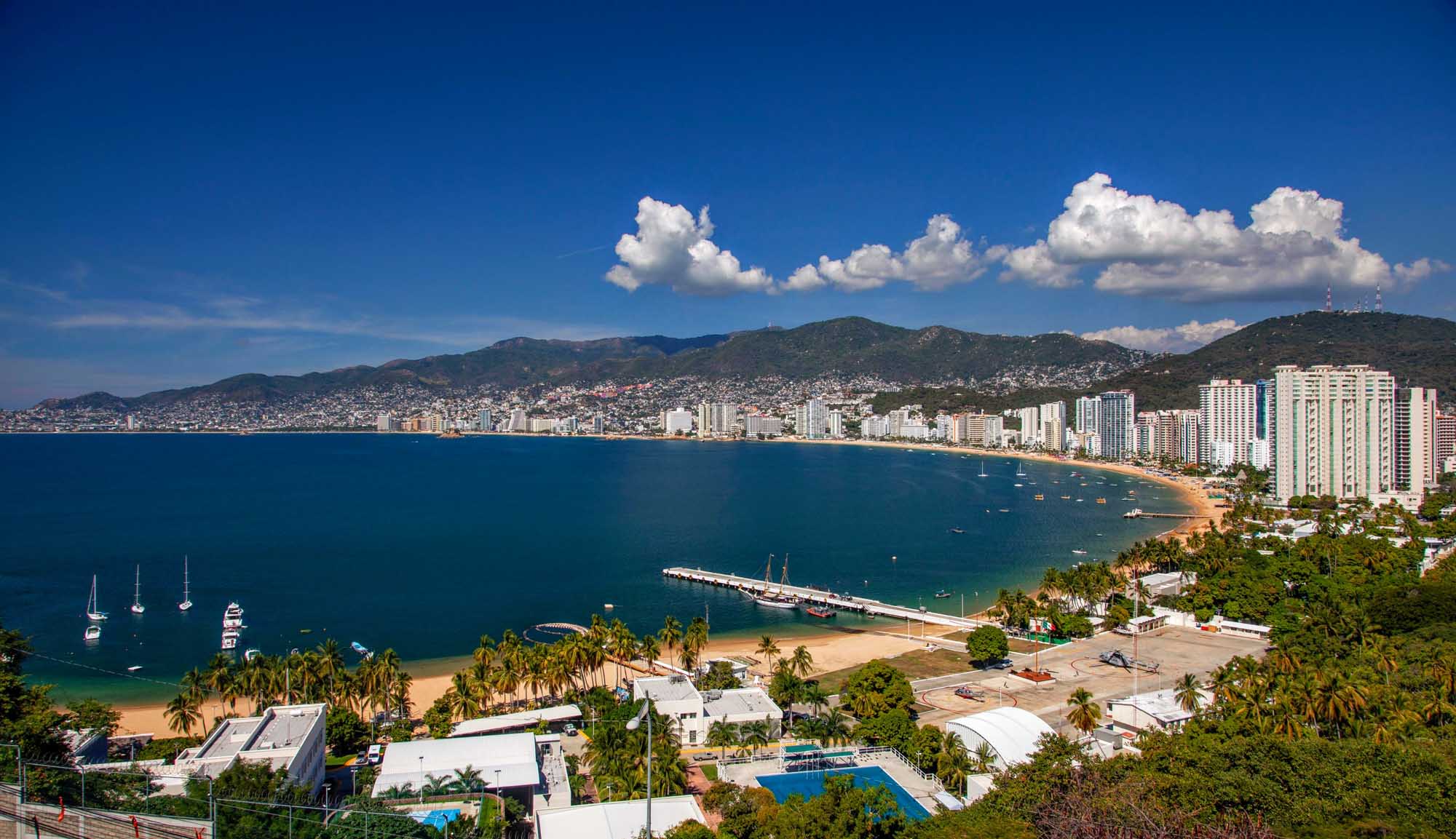 A panoramic view of Acapulco's stunning bay featuring turquoise waters, a curved sandy beach lined with high-rise hotels and resorts, sailboats anchored in the bay, and lush green hills rising behind the city. The scene captures the essence of this famous Mexican beach destination with clear blue skies and puffy white clouds.