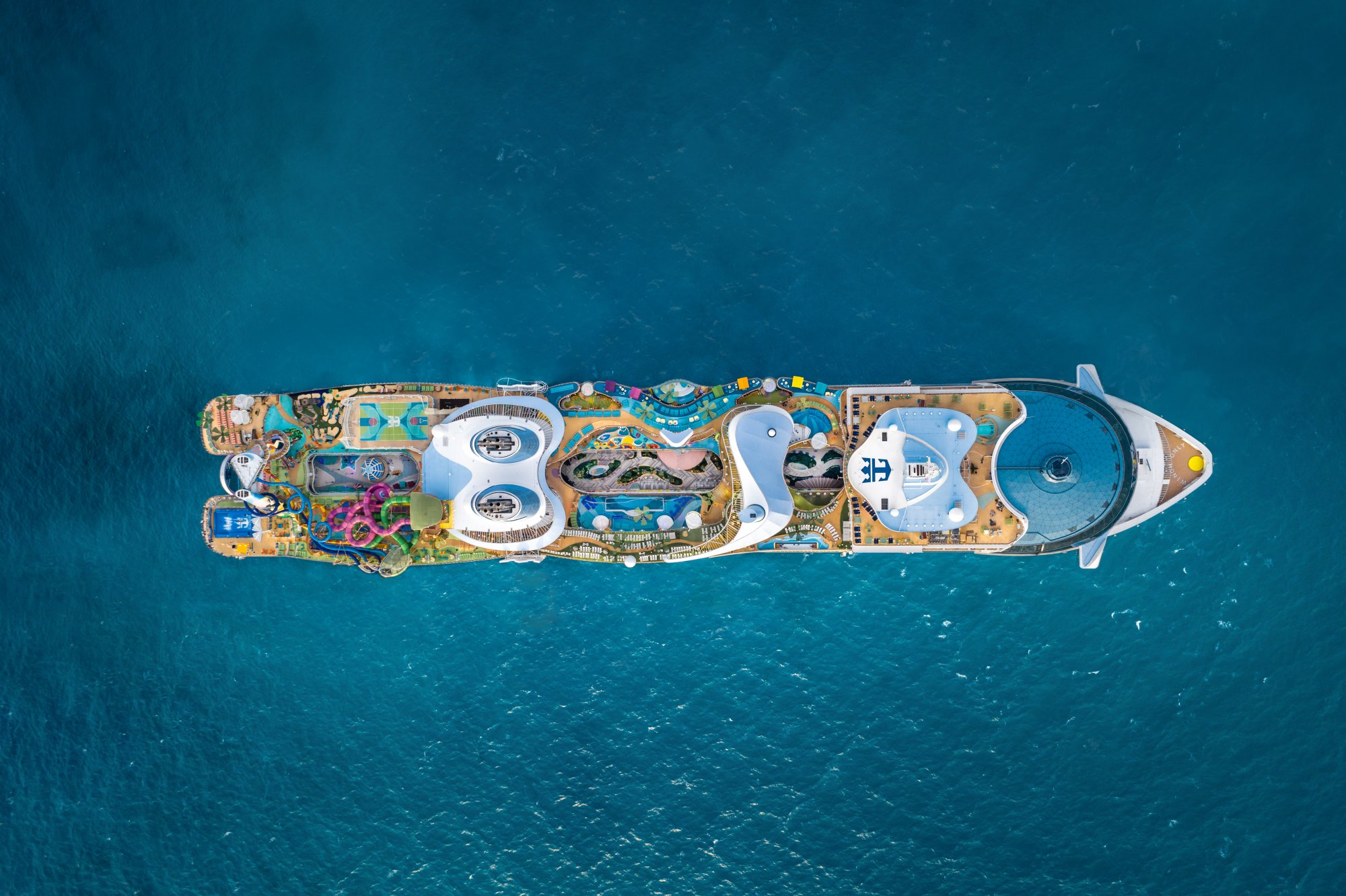 An overhead aerial view of a large modern cruise ship anchored in turquoise Caribbean waters, showcasing the expansive deck layout with multiple pools, recreational areas, and amenities visible from directly above.
