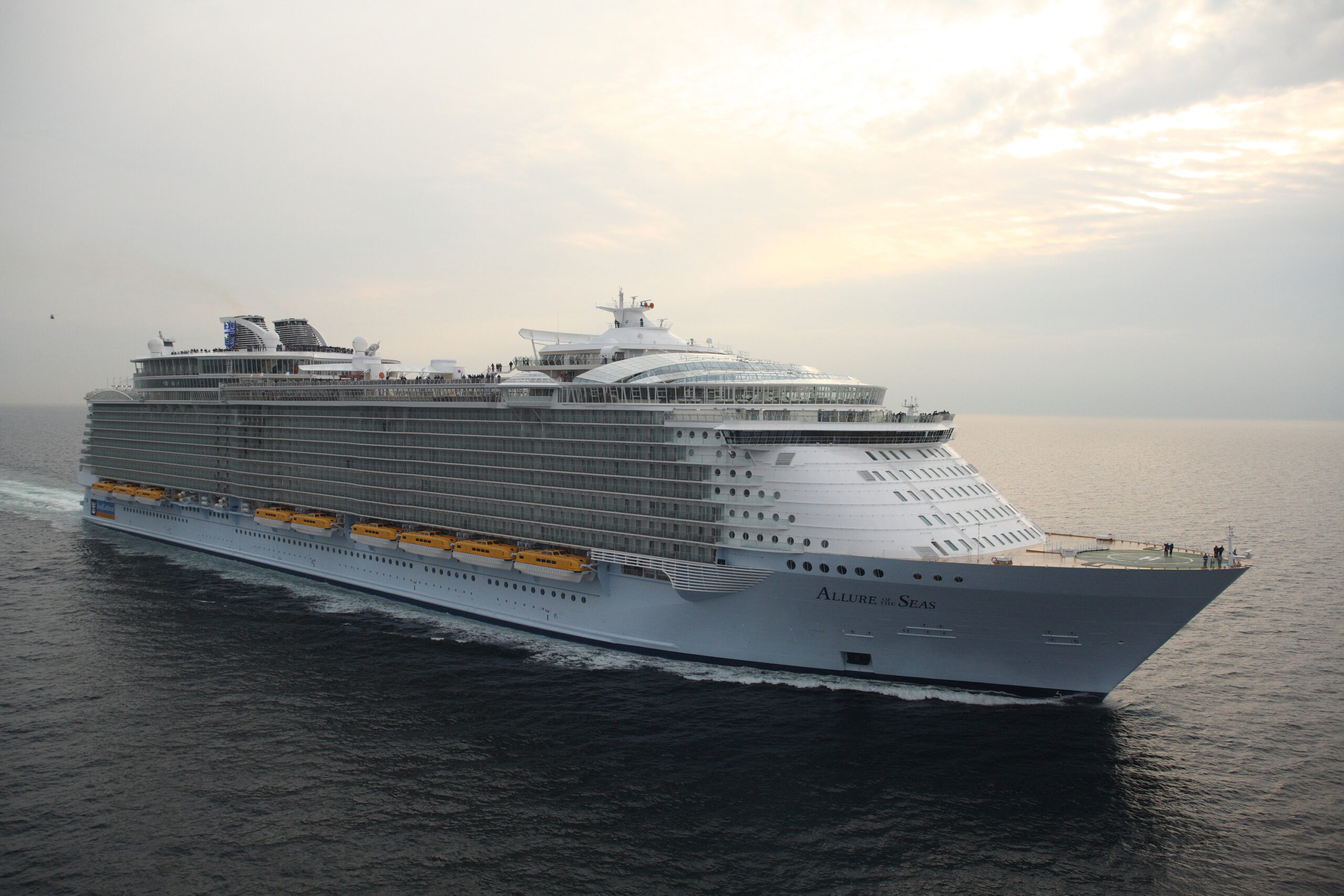 Allure of the Seas Cruise Ship at Sea A massive Royal Caribbean cruise ship sailing on open waters during a soft, overcast day. The Allure of the Seas showcases its impressive size and sleek design with multiple decks and yellow lifeboats.