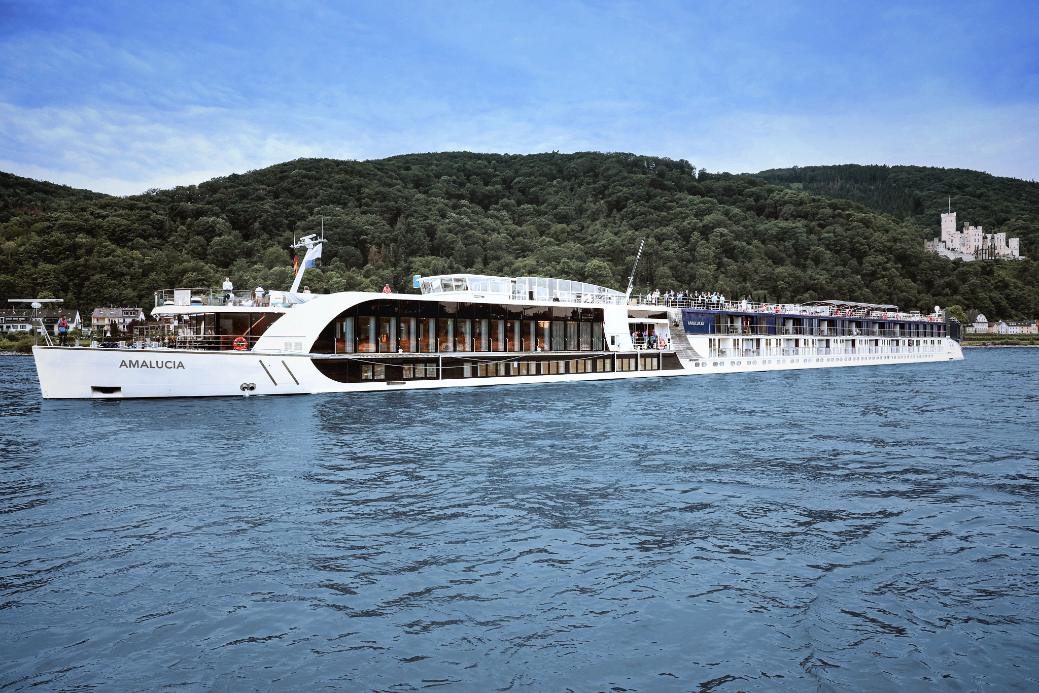 The AmaLucia, a modern river cruise ship, navigates the Rhine River with forested hillsides and a castle visible in the background. The ship features contemporary white hull design with large panoramic windows and multiple passenger decks characteristic of European river cruising.