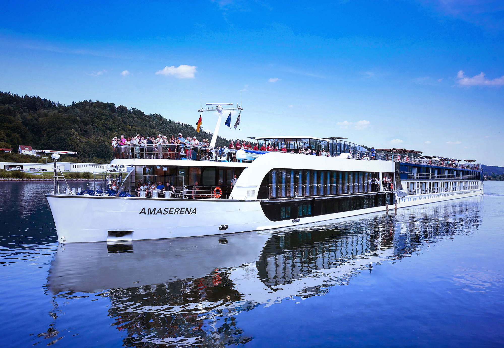 The AmaSerena, a modern river cruise ship, sails through calm blue waters with passengers visible on the upper deck. The vessel features a distinctive white hull with large panoramic windows, multiple decks, and flags displayed from the mast, set against a backdrop of forested hills and clear blue sky.