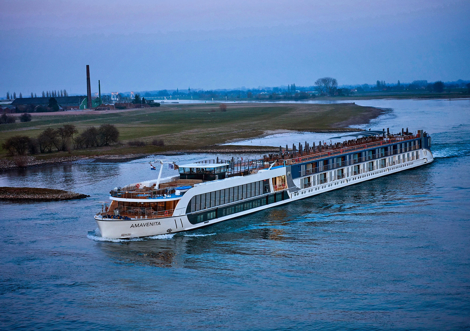 The AmaVenita, a modern river cruise ship, navigates through a wide European river with lush green banks and industrial facilities visible in the distance. The elegant vessel features multiple passenger decks with distinctive blue and white livery, characteristic of contemporary river cruise design.