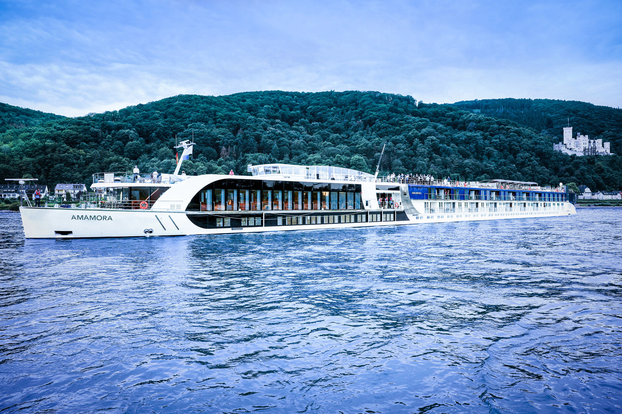 The AmaAmora, a modern river cruise ship, navigates the Rhine River with lush forested hills and a castle visible in the background. The elegant white vessel features contemporary design with large panoramic windows and multiple passenger decks, exemplifying modern river cruising comfort.