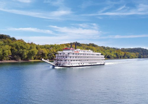 American Splendor Paddlewheel Riverboat on River A classic white paddlewheel riverboat with distinctive red and black smokestacks navigates a calm, blue river lined with lush green forests. The multi-deck vessel represents traditional American river cruising with its iconic paddle wheel design and ornate white superstructure against a scenic natural backdrop.