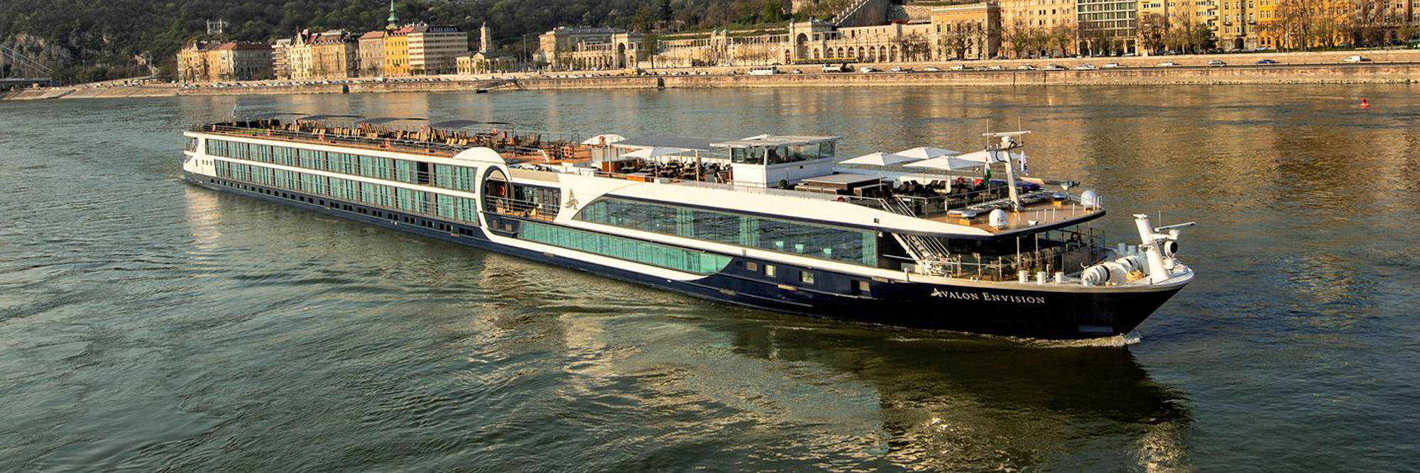 The Avalon Envision, a modern river cruise ship, navigates the Danube River with Budapest's iconic waterfront architecture visible in the background. The vessel features contemporary design with extensive glass paneling, multiple decks, and a sleek white and navy hull, exemplifying modern river cruising luxury.