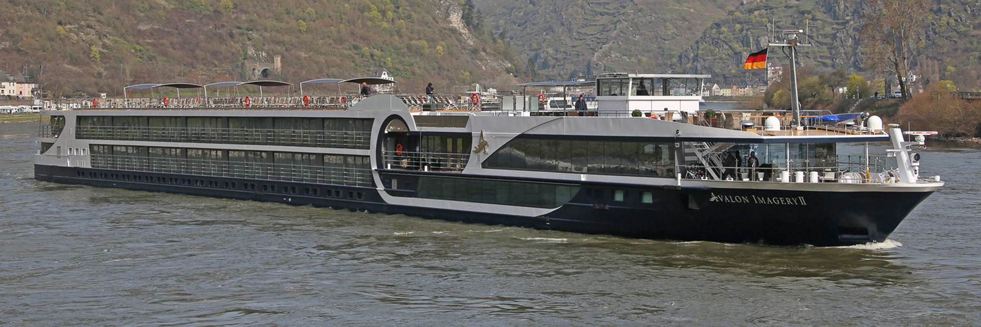 The Avalon Imagery II, a modern river cruise ship, navigates along the Rhine River with forested hillsides and German villages visible in the background. The ship features contemporary design with large panoramic windows, multiple passenger decks, and a German flag, typical of European river cruises.