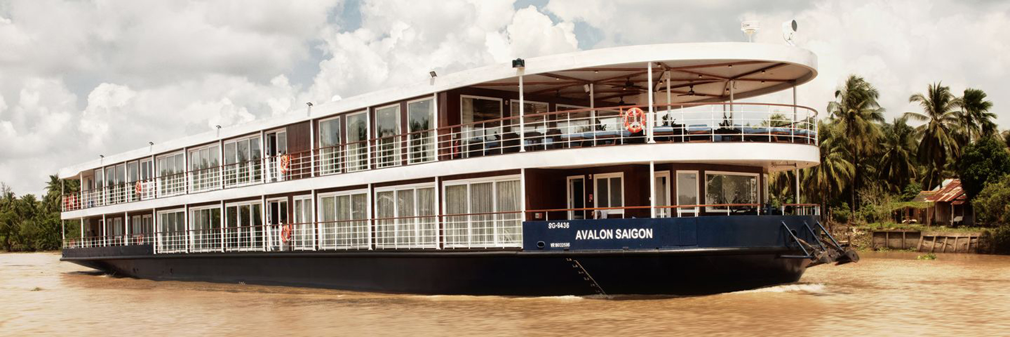 The Avalon Saigon, a modern river cruise ship, is moored along the Mekong River with its distinctive two-deck design featuring expansive windows and an open-air upper deck. The vessel displays contemporary cruise architecture with brown and white exterior coloring, docked near palm-lined riverbanks in Vietnam.
