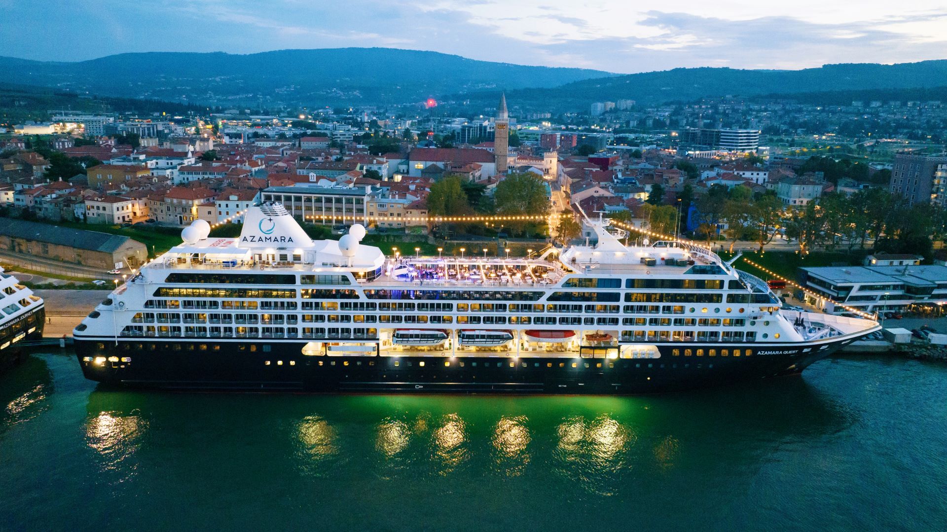 The Azamara Quest luxury cruise ship is illuminated at night while docked in Koper, Slovenia, with the historic city's architecture and church spire visible in the background. The vessel's elegant white superstructure and extensive lighting showcase the ship against the twilight sky and surrounding Mediterranean-style townscape nestled between forested hills.