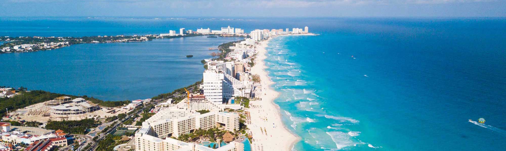 An aerial photograph of Cancun's iconic Hotel Zone showing a dense strip of high-rise beachfront resorts lining a pristine white sand beach with turquoise Caribbean waters. The image captures the lagoon on the left, the developed beachfront on a narrow peninsula, and the open Caribbean Sea on the right, with numerous tourists visible on the beach.