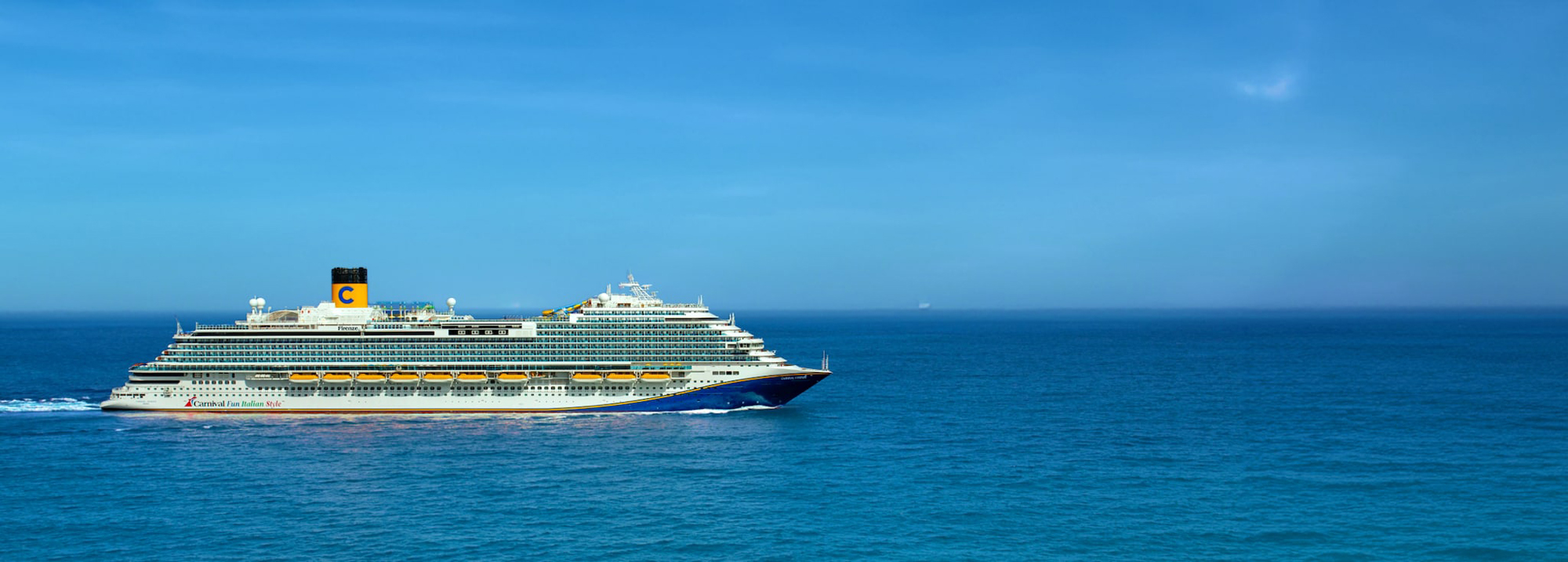 A large modern cruise ship with distinctive yellow and blue funnel livery sails through calm, deep blue ocean waters under clear skies. The vessel displays the characteristic Carnival Cruise Line branding with the 'C' logo prominently visible on its funnel.