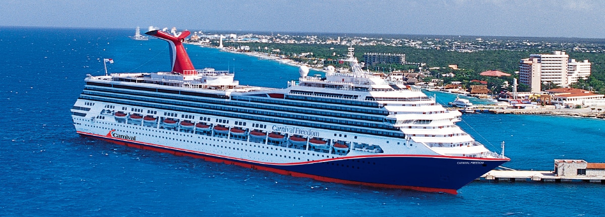 The Carnival Freedom, a large cruise ship featuring the distinctive red funnel and blue hull livery of Carnival Cruise Line, is anchored in crystal-clear turquoise waters at a tropical Caribbean destination. The ship's white superstructure contrasts sharply against the brilliant blue ocean, with a lush, developed coastline visible in the background.