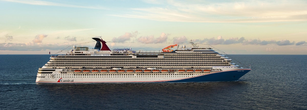 A large modern cruise ship with distinctive Carnival branding and livery sails through calm ocean waters under a partly cloudy sky. The vessel features multiple decks, a prominent red funnel, and the characteristic blue hull typical of contemporary mega-ships.