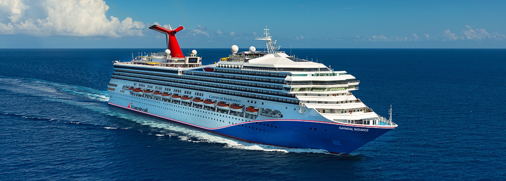An aerial view of the Carnival Radiance, a large modern cruise ship, sailing through deep blue ocean waters under a clear sky with scattered white clouds. The ship features the iconic red Carnival funnel and distinctive blue hull, showcasing multiple passenger decks and amenities.