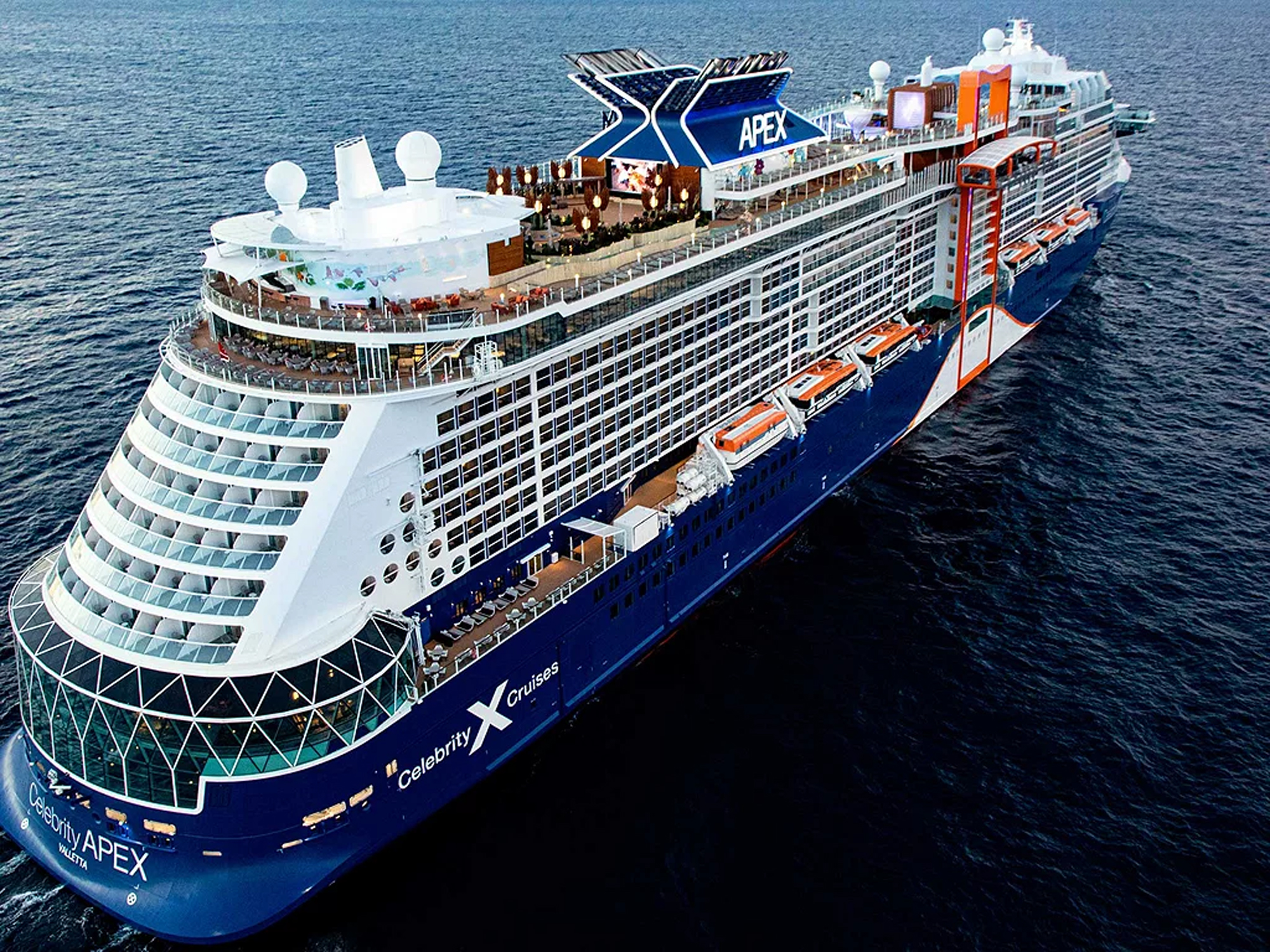 An aerial view of the Celebrity Apex, a modern mega-ship operated by Celebrity Cruises, showcasing its distinctive design with the characteristic 'X' logo on the funnel, white superstructure, blue hull, and orange lifeboats. The ship displays the cruise line's signature branding and contemporary architecture on the open ocean.