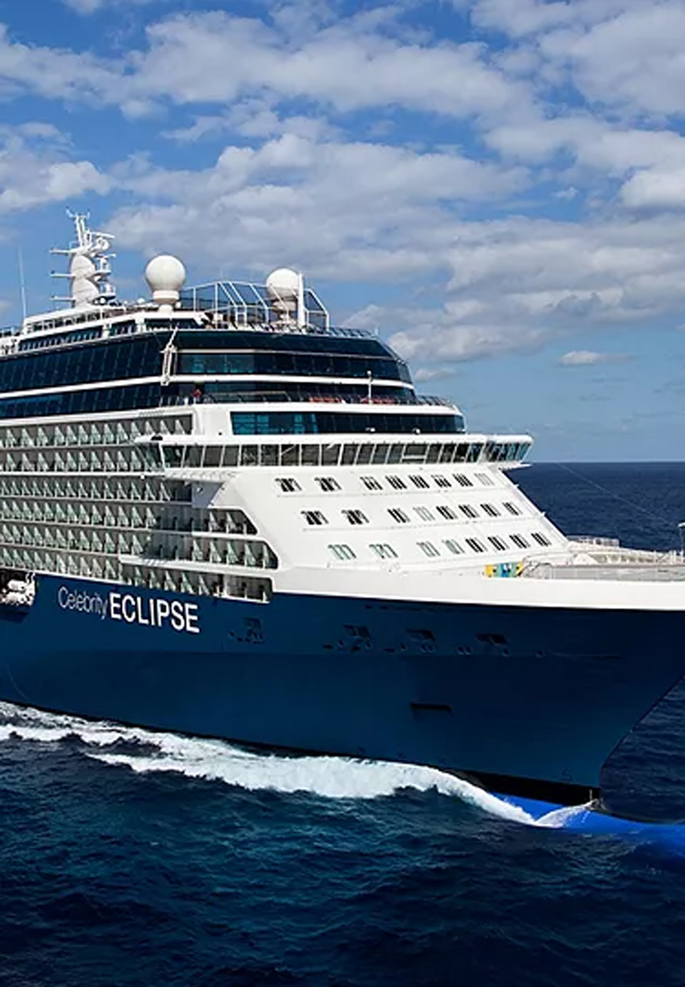 The Celebrity Eclipse, a modern luxury cruise ship, is docked at port with its distinctive dark blue hull and white superstructure visible against a bright blue sky with white clouds. The ship displays multiple decks with rows of cabin windows and sophisticated maritime infrastructure including communication domes on the upper deck.