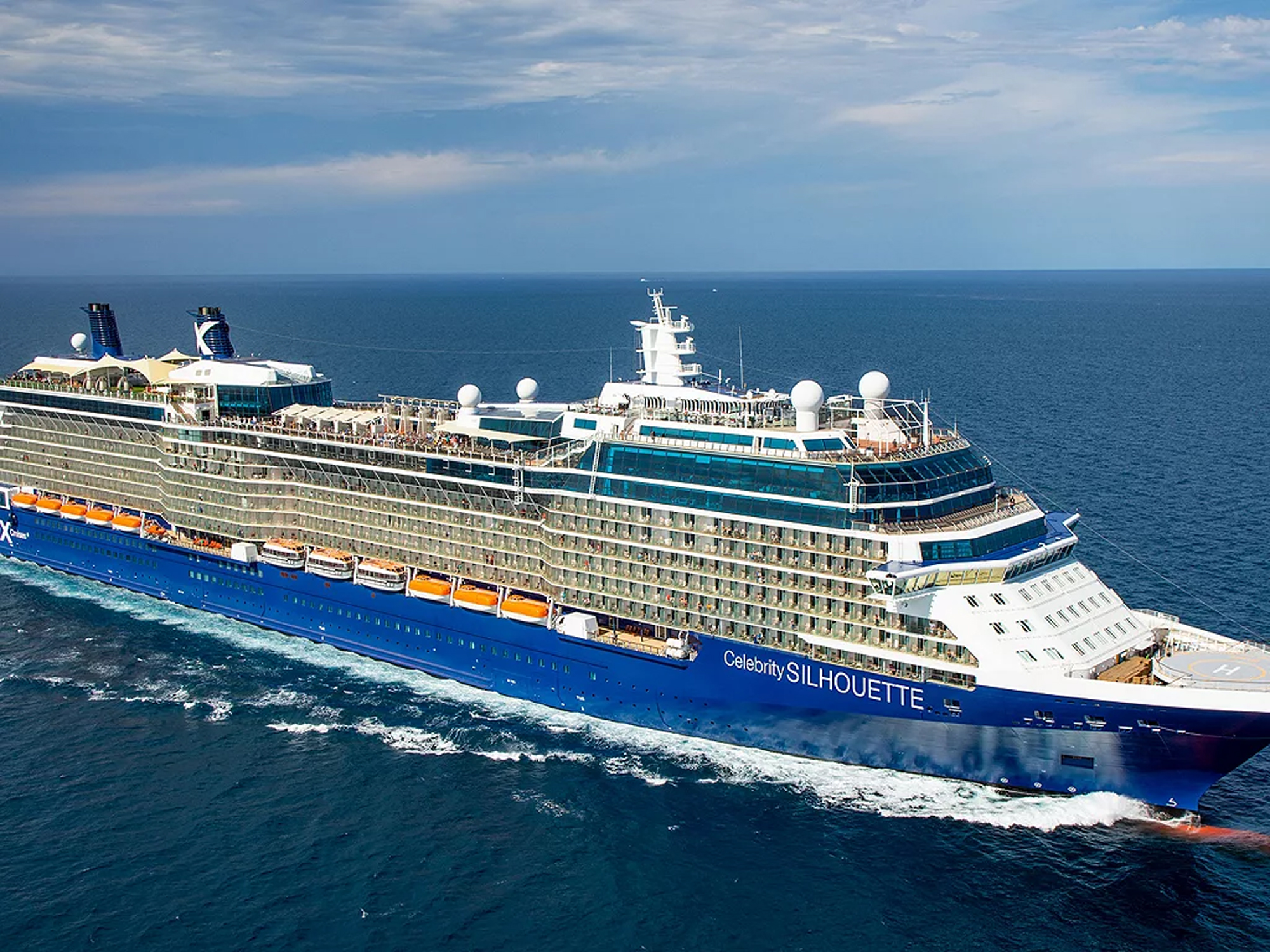 A large modern cruise ship, the Celebrity Silhouette, navigates through blue ocean waters with white wave wake visible along its hull. The vessel features a distinctive blue hull with multiple decks, white superstructure, and modern maritime technology visible on the bridge.