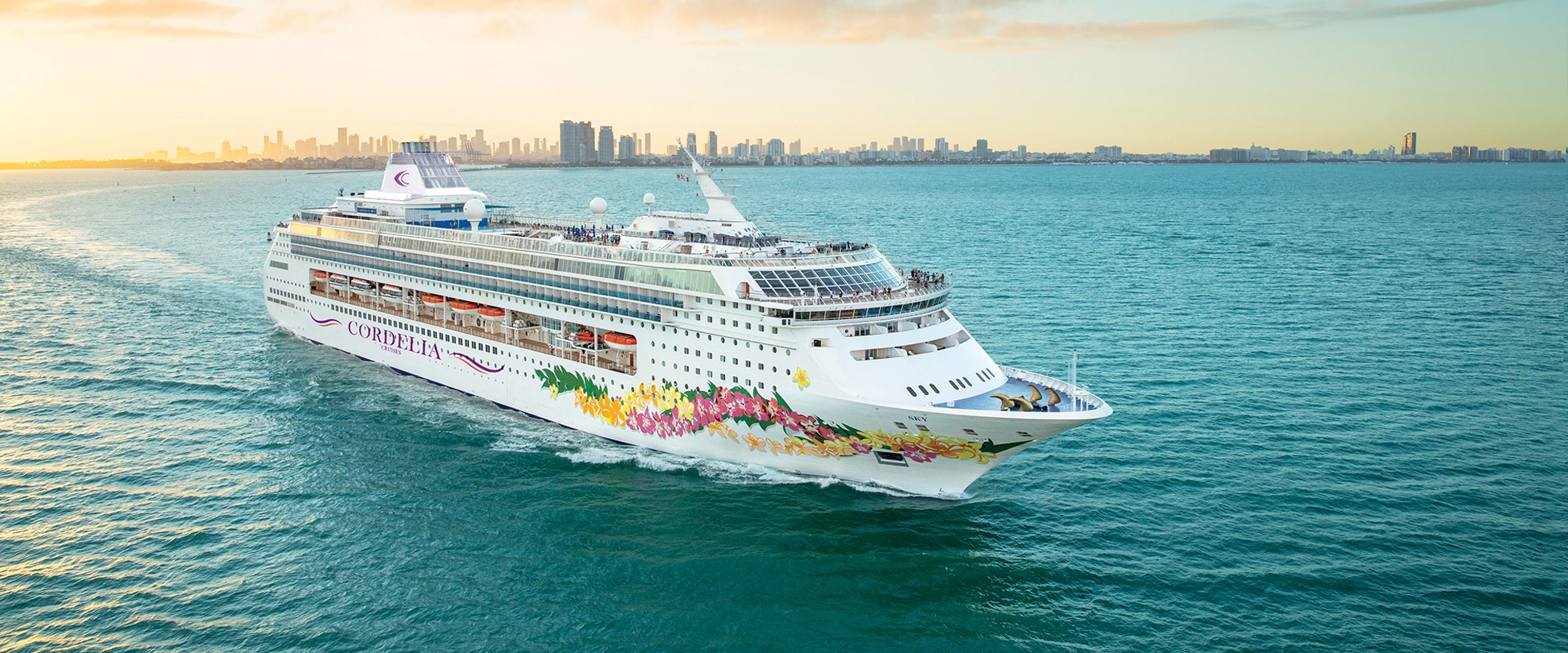 Cruise Ship Departing Miami Waters at Sunset A large modern cruise ship with vibrant floral artwork on its hull sails through turquoise waters off Miami, with the city's skyline visible in the golden evening light. The vessel appears to be departing or arriving at port, captured from an aerial perspective.