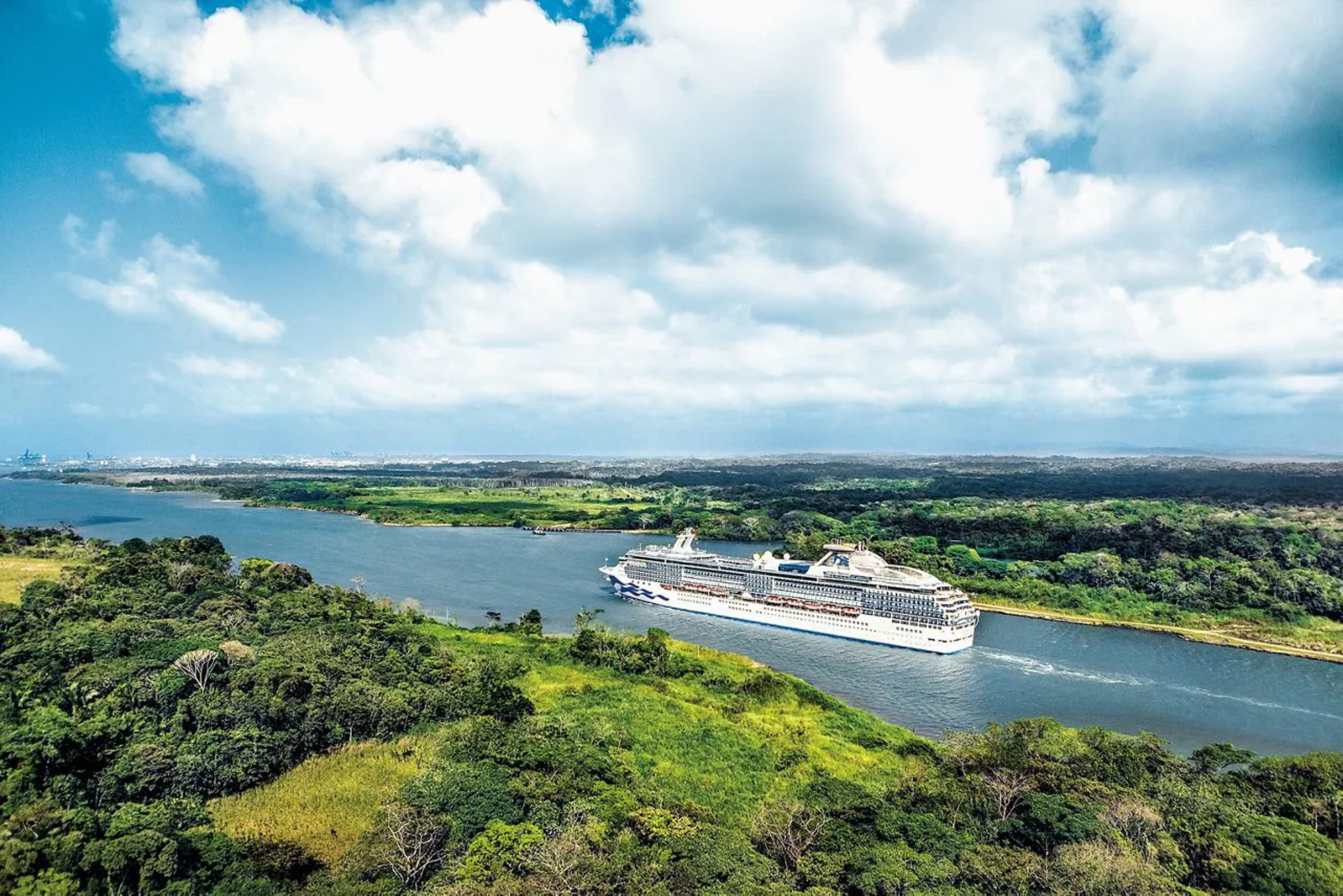 Cruise Ship Transiting Gatun Lake Canal A large modern cruise ship navigates through Gatun Lake surrounded by lush tropical rainforest and verdant vegetation. The image captures an aerial perspective of the vessel in the waterway with dense green foliage on both shorelines and a bright blue sky with white clouds overhead.